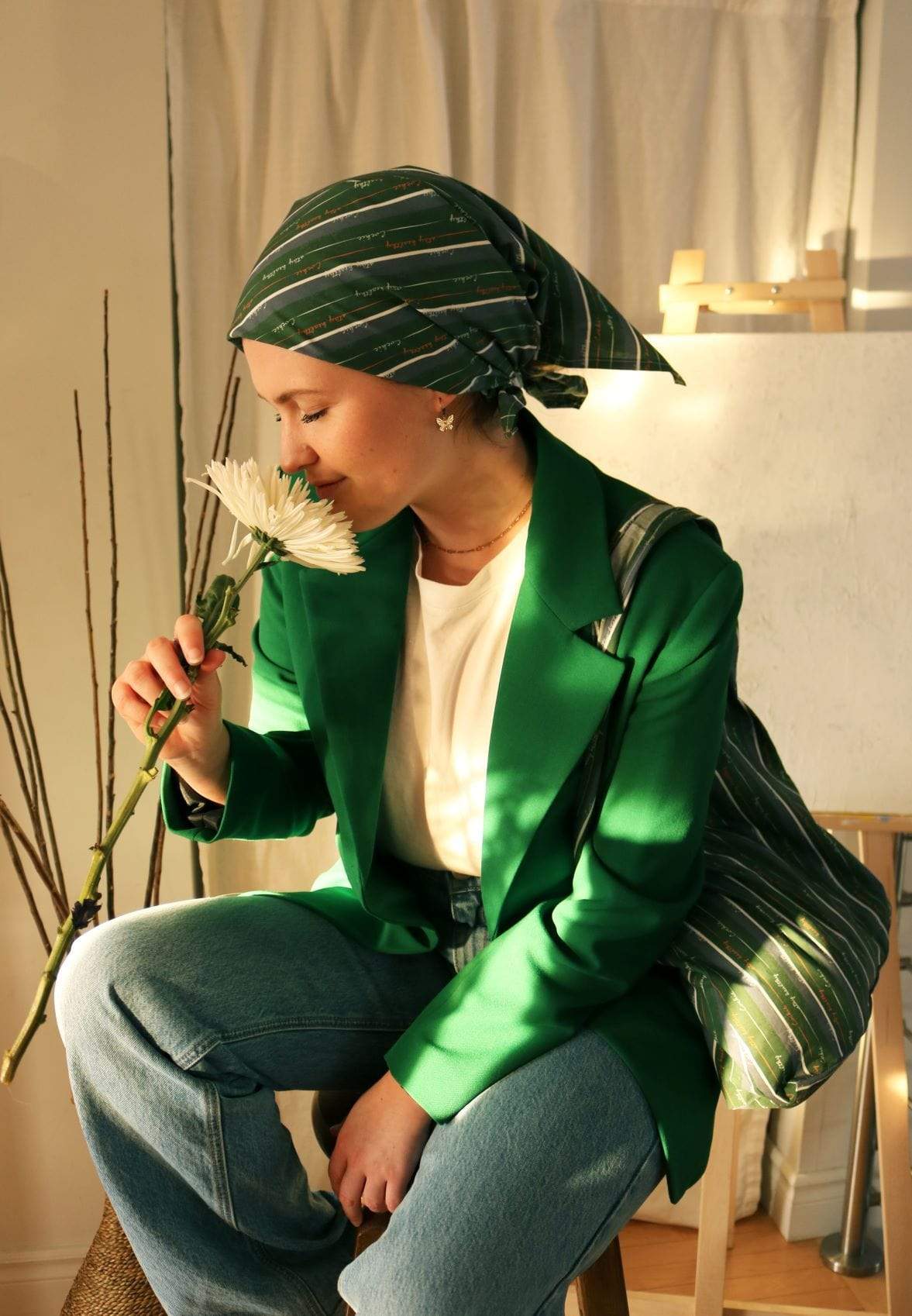 Elegant woman sitting indoors, wearing a green blazer, matching headscarf, and carrying a stylish striped green bag, gently smelling a white flower, bathed in warm natural light with a minimalist background.